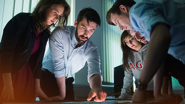 Zdjęcie studentów w ciemnym pomieszczeniu, pochylających się nad stołem z ekranem dotykowym. Jeden z mężczyzn wskazuje palcem na ekran.