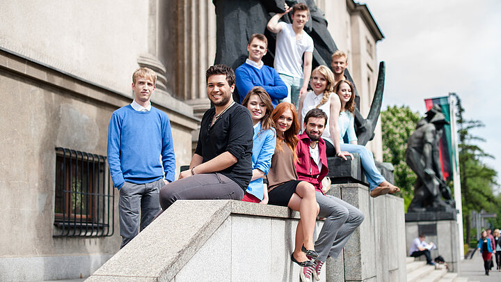 Grupa młodych, uśmiechniętych ludzi na schodach przed dużym budynkiem. 